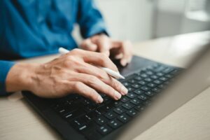 Close-up of businessperson hands typing on laptop keyboard while holding stylus pen in home office. Concept of freelance work, job search, digital communication, and modern technology workspace.